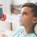 Boy lying in bed wearing a nebulizer mask connected to a handheld inhaler held by an adult caregiver for breathing treatment.