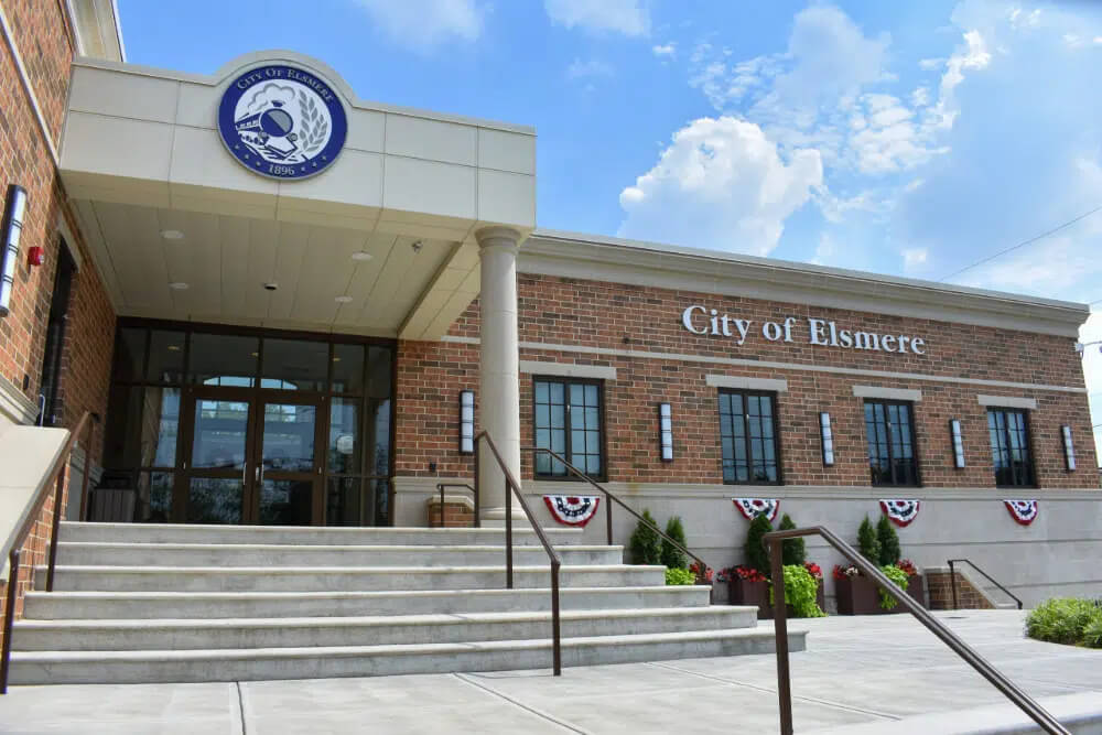 Elsmere | Pediatrics Of Florence City of Elsmere municipal building with brick facade, glass doors, and stairs leading to the entrance, decorated with red, white, and blue bunting.