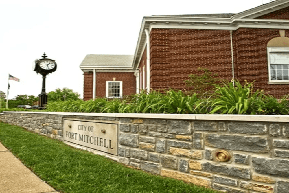 Fort Mitchell | Pediatrics Of Florence Stone retaining wall with a plaque reading 'City of Fort Mitchell' in front of a brick municipal building and a vintage clock tower nearby, with greenery around.
