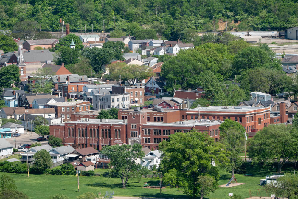 Aerial view of a small town with a large red brick school building in the foreground, surrounded by houses and green trees.