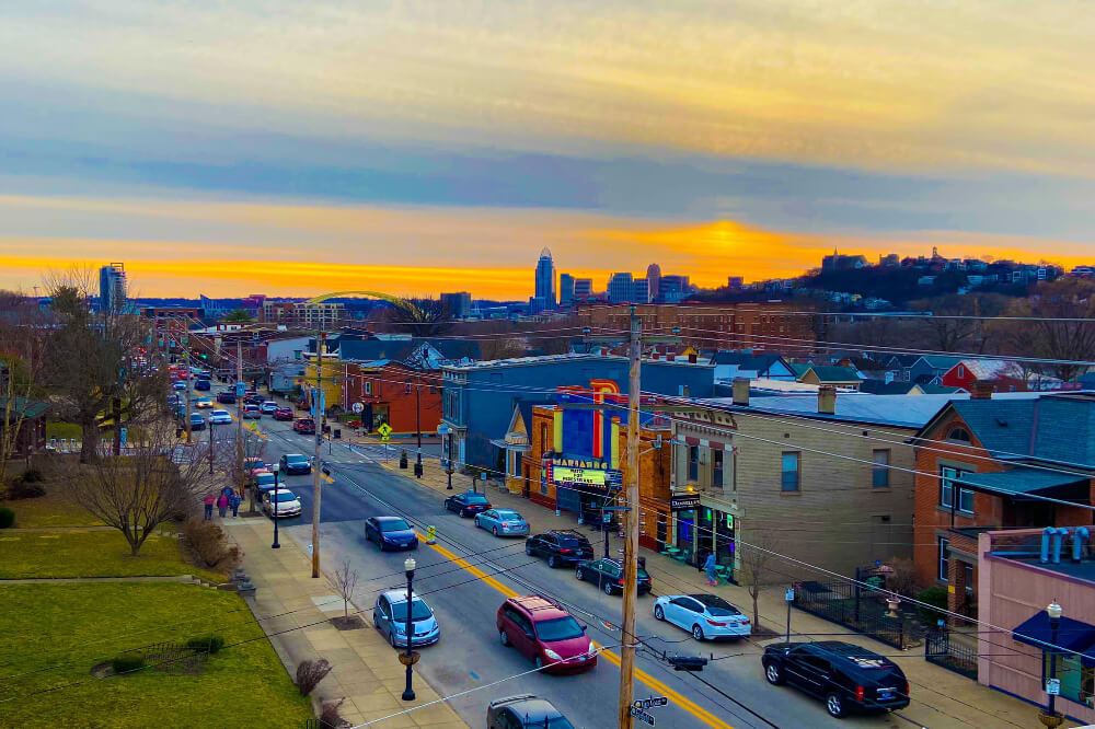 Bellevue KY | Pediatrics Of Florence Sunset over a busy city street with cars, storefronts, and a distant skyline.