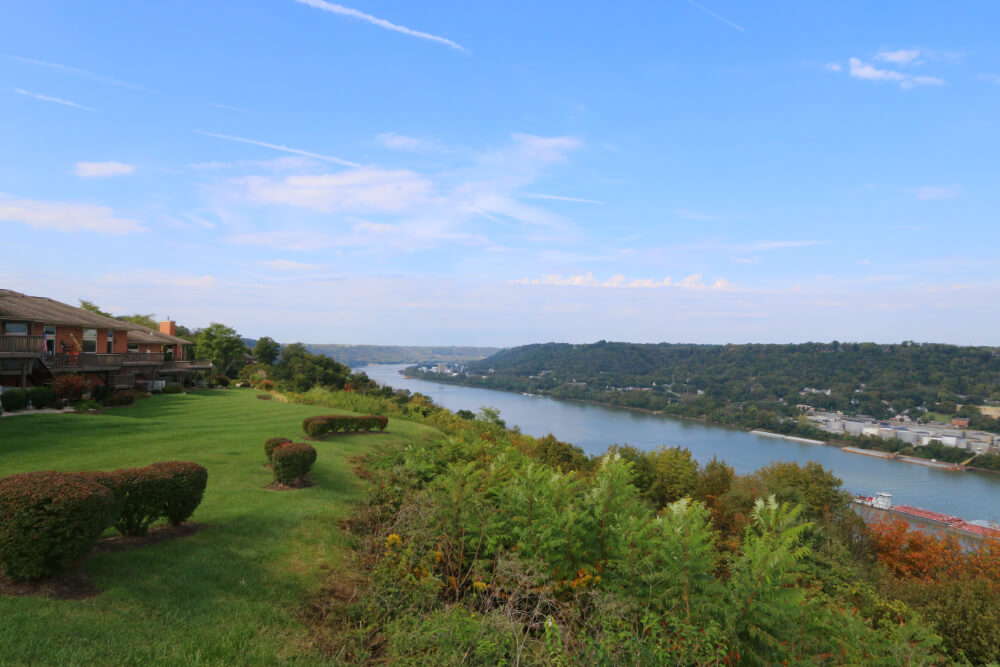 Villa Hills | Pediatrics Of Florence Scenic overlook of a river valley with a green lawn, trimmed hedges, and houses to the left under a blue sky.